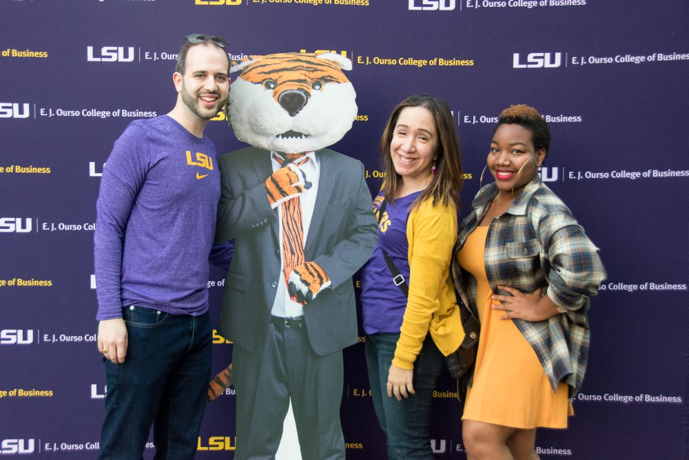 One man and two women wearing purple and gold smile in front of a colleg of business backdrop.