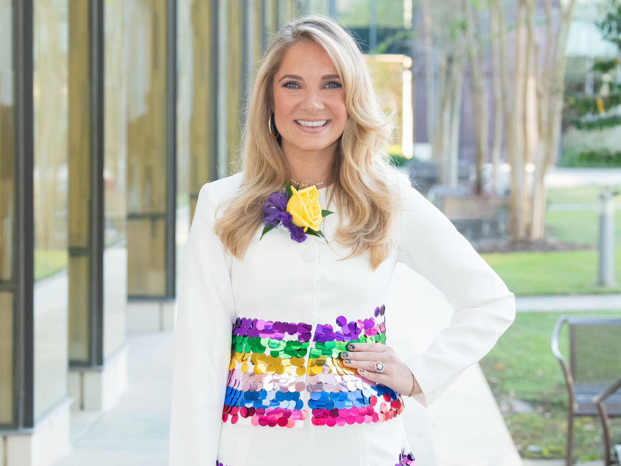 Jaime Glas is in a white dress with colorful sequines. She is standing outside with trees and columns in the background.